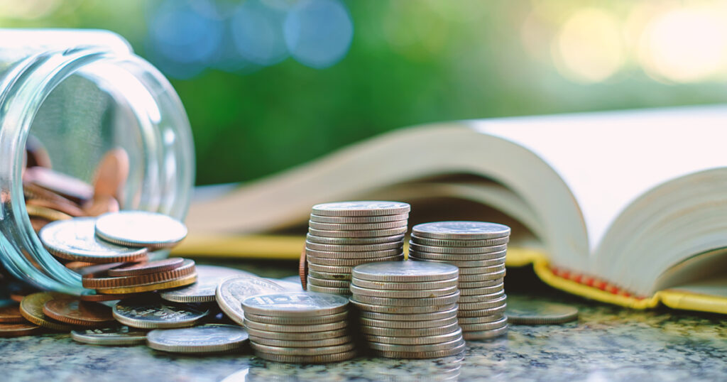 Pile of money coins in and outside the glass jar on blurred book and natural green background for financial and education concept