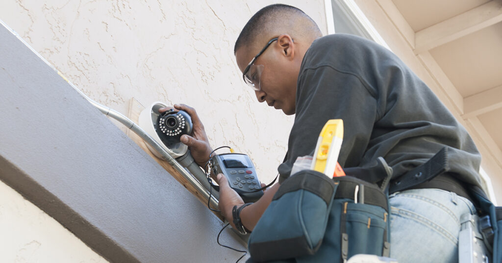 A technician uses a monitor to adjust a security camera while installing the system to the exterior of a building.